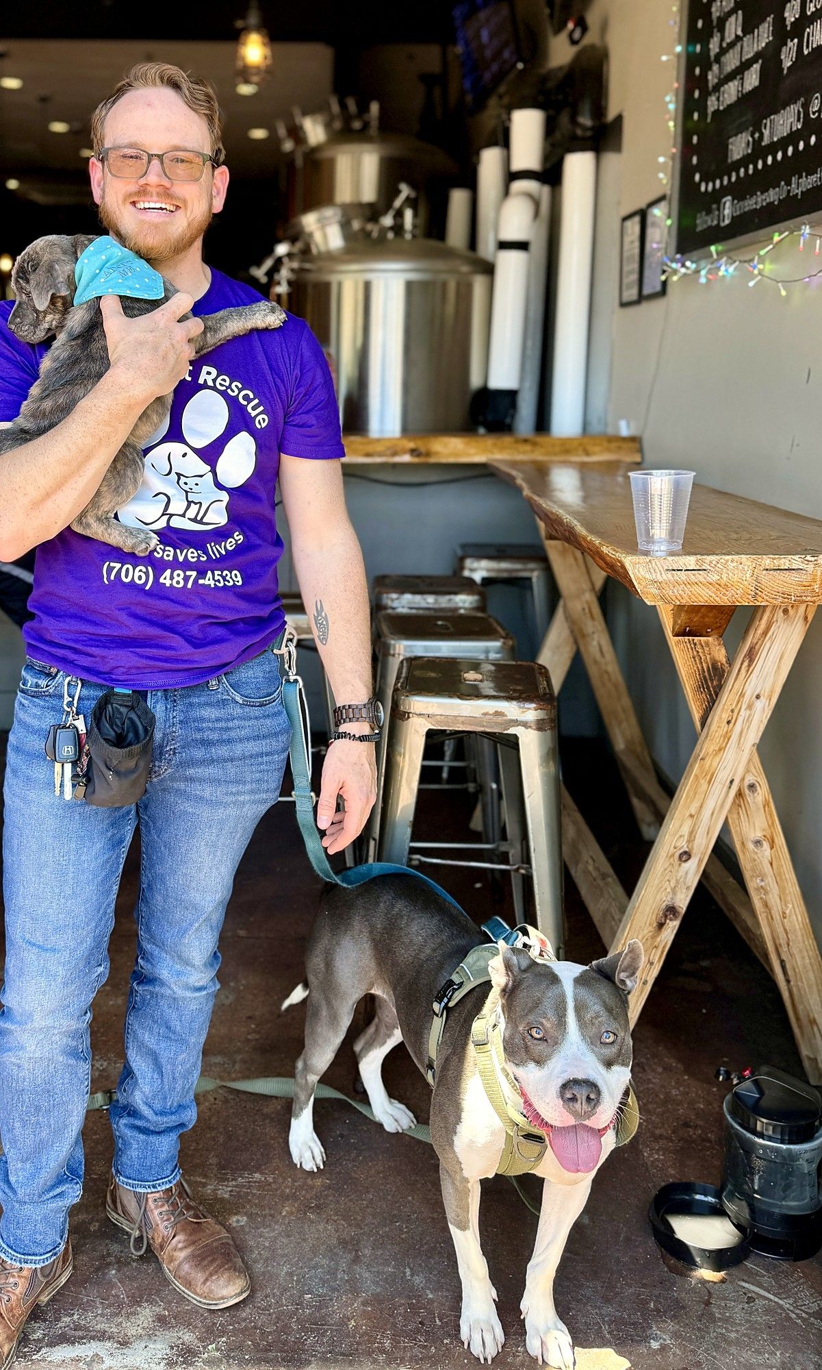 Phillip in a rescue volunteer shirt holding a puppy, with Lucky standing at his feet in a taproom